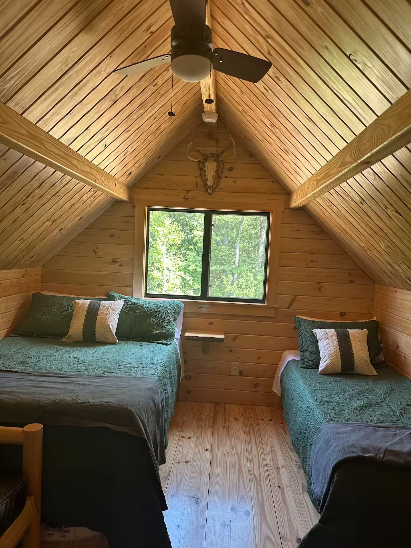 Una cabaña con dos camas, un ventilador de techo y un ventanal con vistas a árboles.