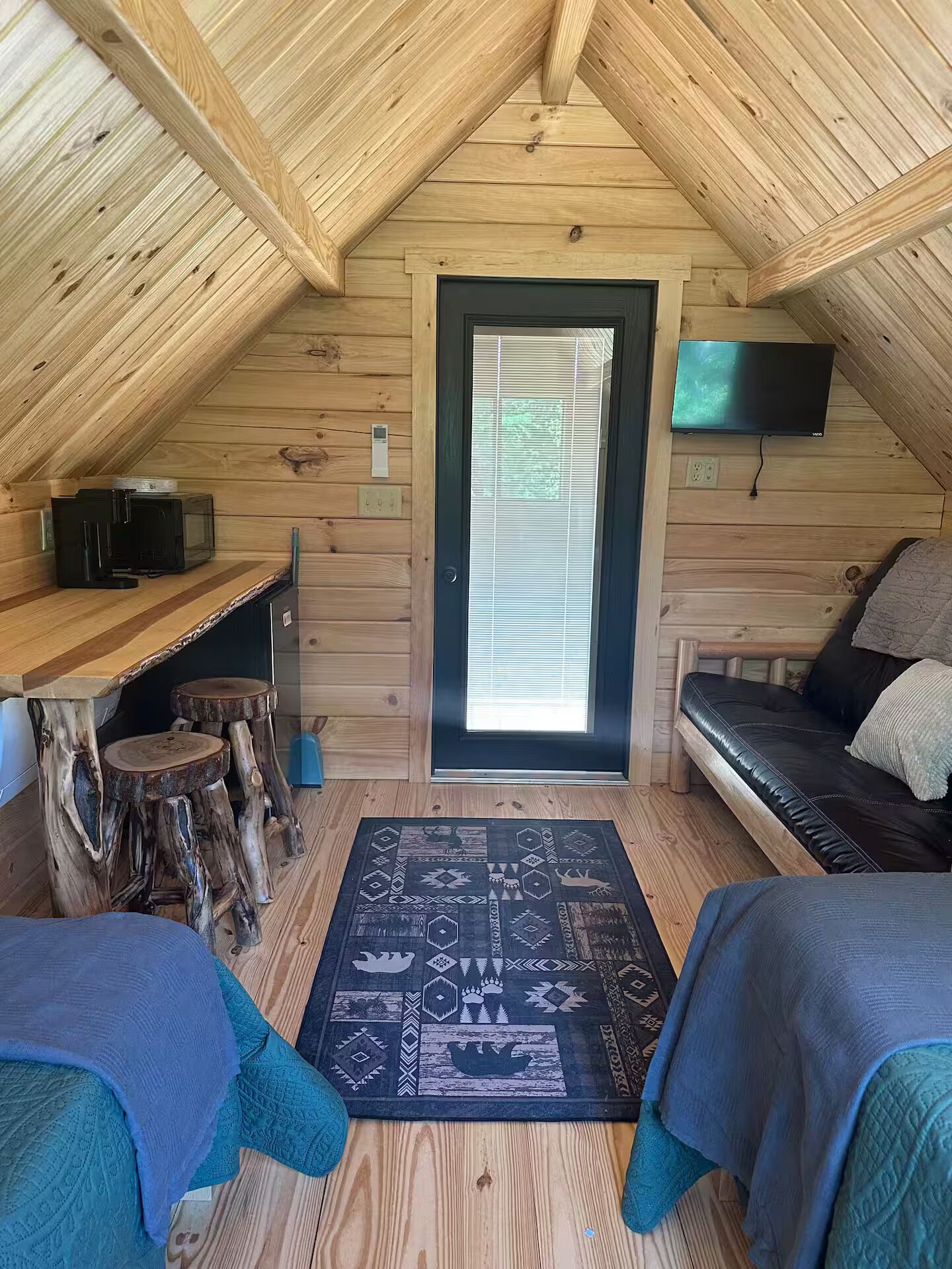 A cozy attic room with a sloped ceiling, a television mounted on the wall, a window with blinds, a wooden desk, a couch, and a patterned rug.