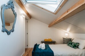 Ein Schlafzimmer mit Dachfenster, einem Bett mit blauer Decke, einem Nachttisch mit Lampe und einem Spiegel an der Wand.