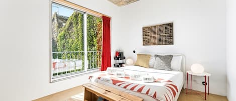 A modern bedroom with a large window, a bed with a white headboard, a bedside table, and a small side table with a lamp.