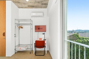 Une chambre d’hôtel avec un étagère en métal, une chaise rouge, un téléviseur à écran plat et une vue sur le paysage urbain.