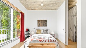 A modern bedroom with a large bed, a wooden bench, a bookshelf, and a view of greenery through a large window. - GuestReady - House of Many Faces - Room 2 (Porto)