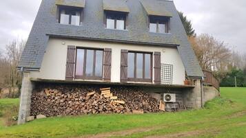 Ett hus med ett sluttande tak, tre fönster med fönsterluckor, en hög med ved och en trädgård.