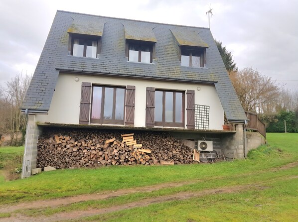 Une maison avec un toit en pente, trois fenêtres avec volets, une pile de bois de chauffage et un jardin.