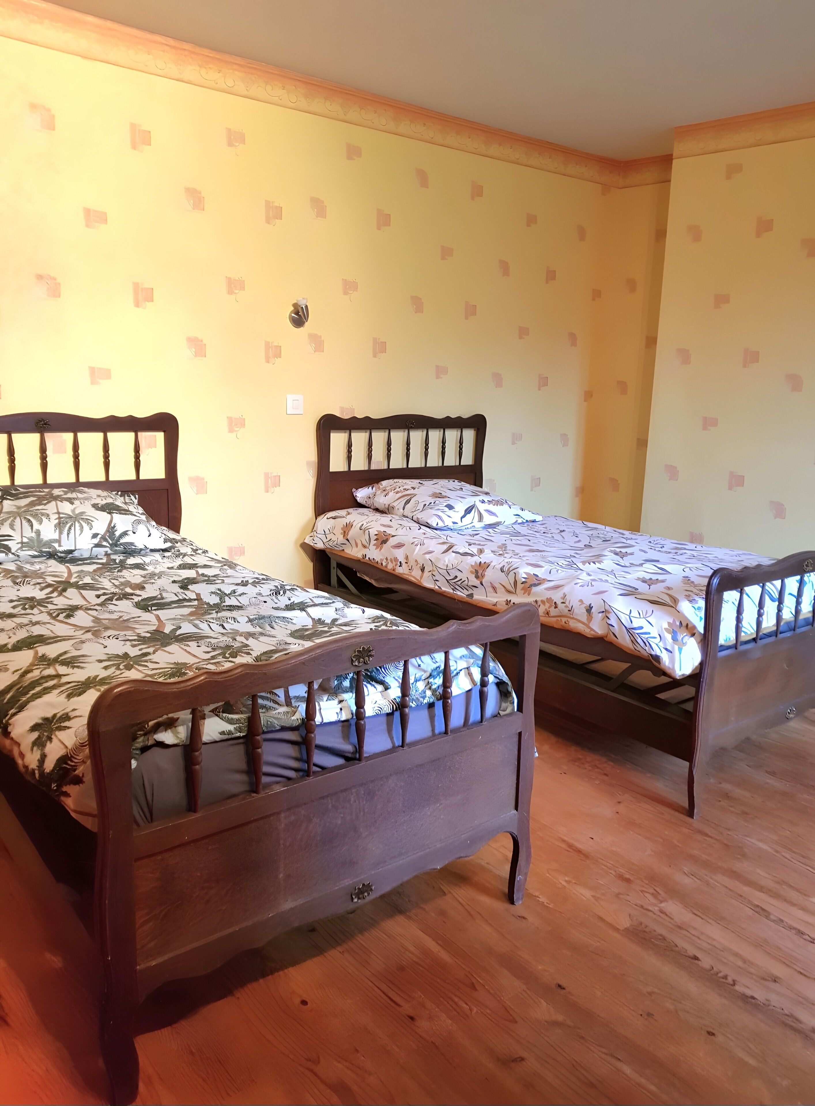 Une chambre avec deux lits en bois, chacun doté d’une tête de lit et d’un pied de lit, et recouverte d’une couvre-lit à motifs. Les murs sont d’une couleur jaune pâle, avec un motif discret.