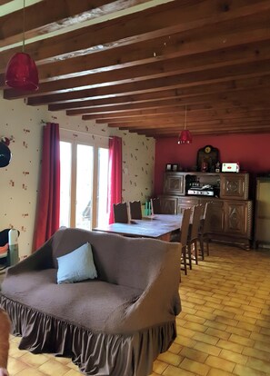 Un salon chaleureux avec un canapé, une table à manger et des chaises, des poutres en bois au plafond et un coin cuisine équipé de placards et d’appareils électroménagers.