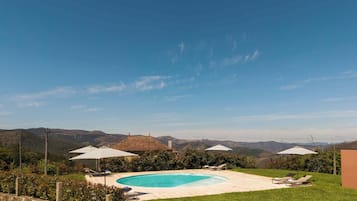 A swimming pool with lounge chairs and umbrellas, surrounded by a stone wall and greenery, with mountains in the background.