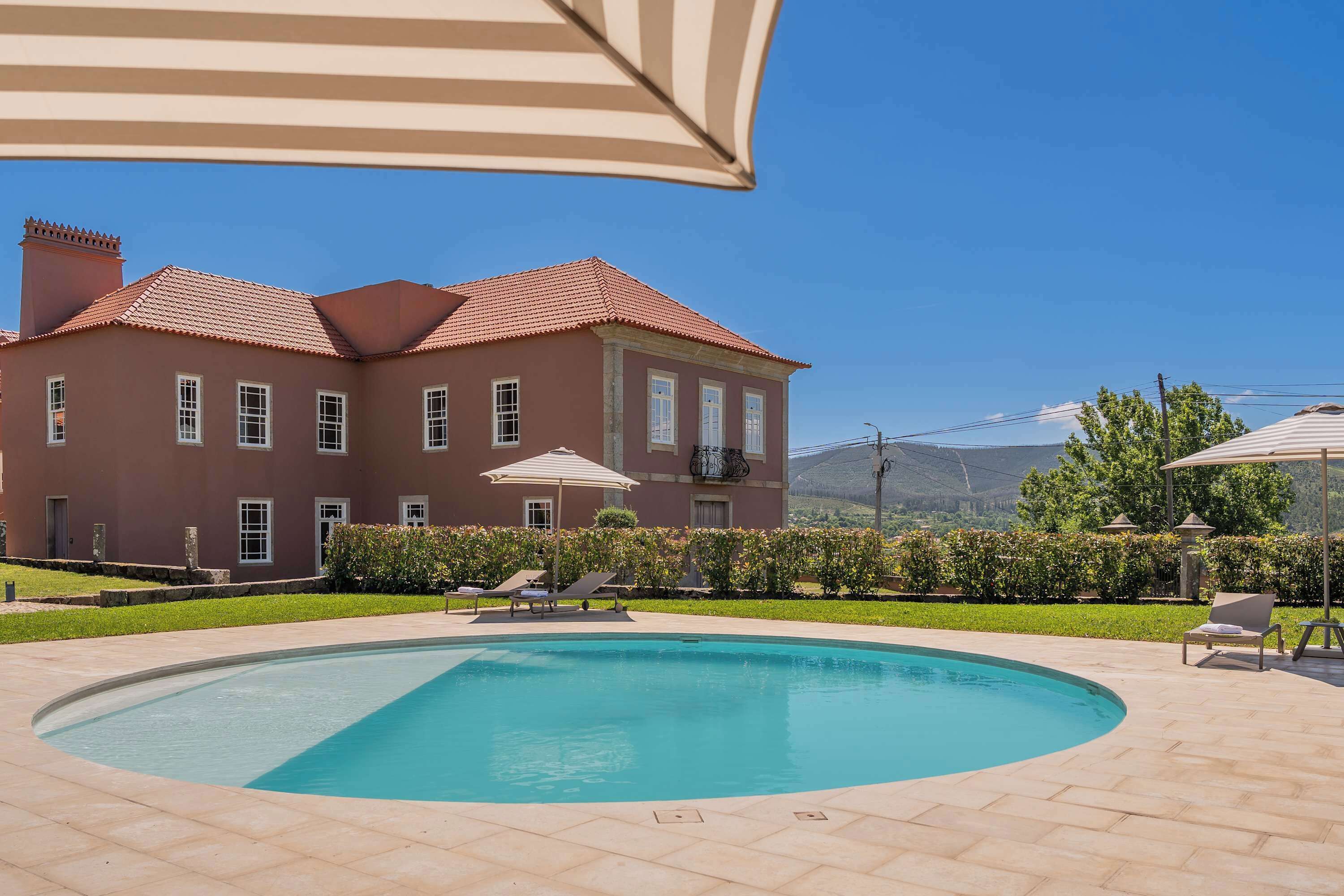 Une piscine avec des chaises longues et des parasols, une grande maison avec un toit rouge et un ciel bleu azur.
