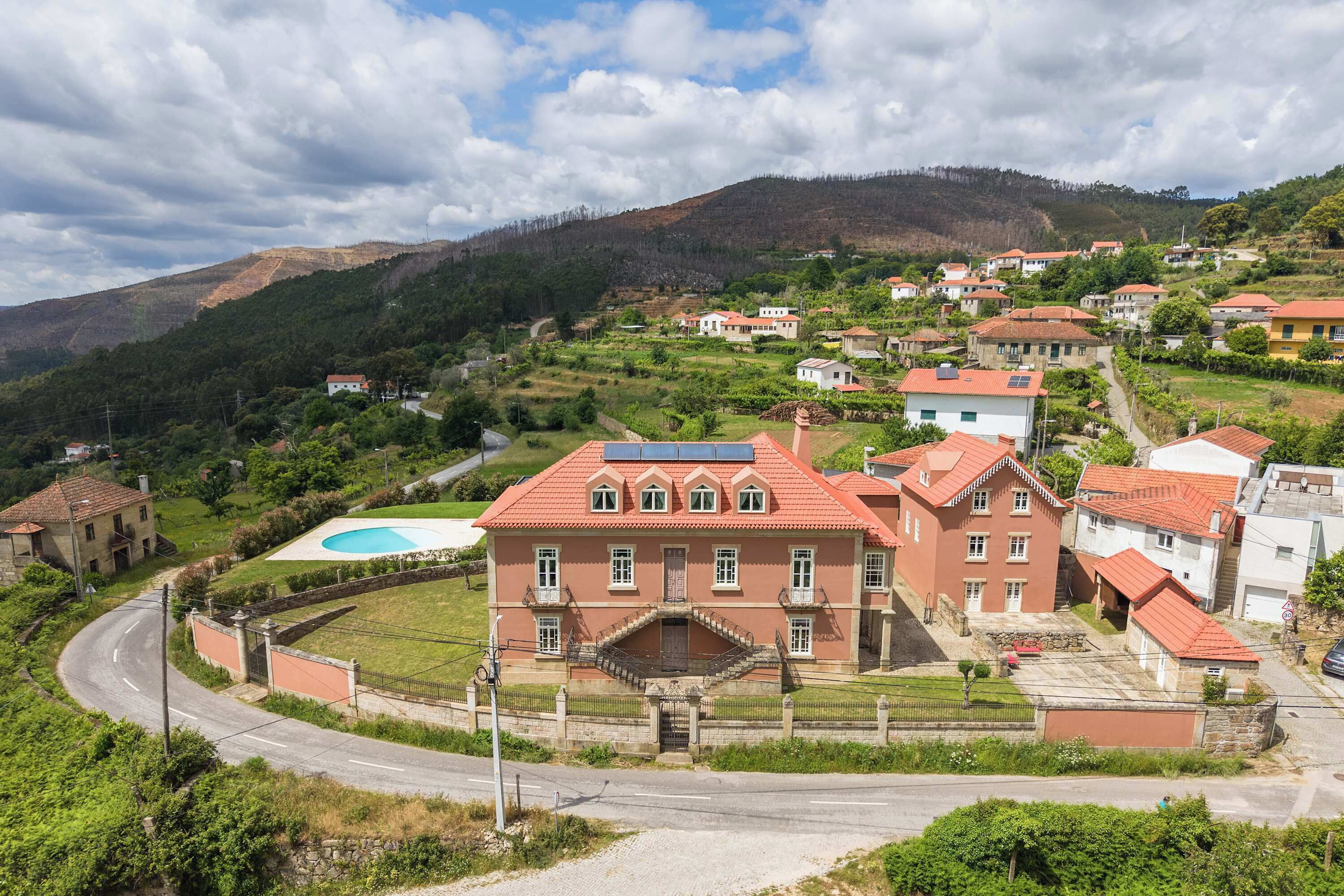 Un edificio grande de techo rojo con una escalera central, rodeado de casas más pequeñas, ubicado en una zona montañosa con cielo despejado.