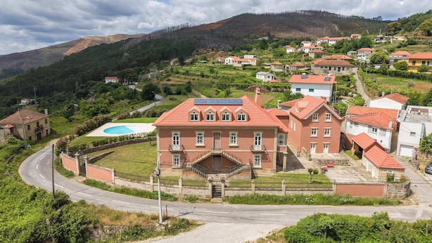 A large, red-roofed building with a central staircase, surrounded by smaller houses, set in a hilly area with a clear sky.