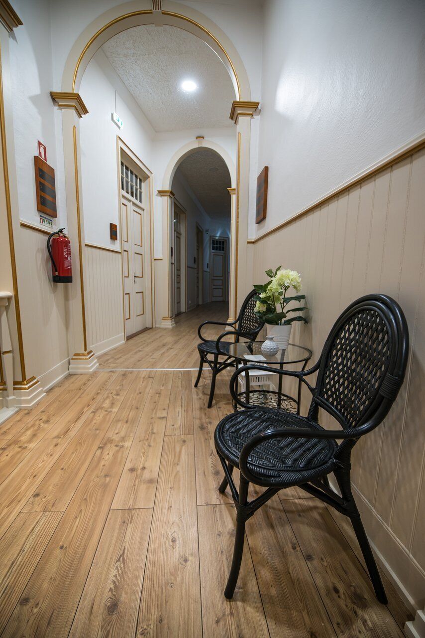 Un couloir doté d’un plancher en bois, de chaises en osier noir, d’une petite table sur laquelle se trouve un vase, et d’un extincteur fixé au mur.