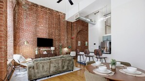 Ein modernes Wohnzimmer mit Backsteinwand, einer Couch, einem Esstisch für vier Personen und einer Küchenzeile.