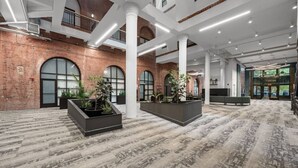 Eine geräumige Lobby mit hohen Decken, großen Fenstern und einer Backsteinwand. Es gibt mehrere Sitzbereiche und Pflanzgefäße mit Grünpflanzen.