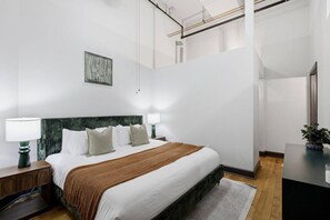 Ett modernt hotellrum med en stor säng, två sängbord, ett litet bord, ett skrivbord, en stol, en spegel och en krukväxt.