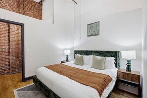 A hotel room with a large bed, two bedside tables with lamps, a brick wall, and a framed artwork on the wall. - Luxury Loft + King Bed Heart of Downtown STL (St. Louis)