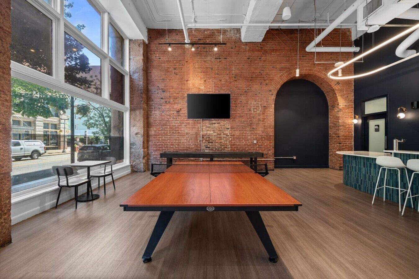 Une table de billard dans une pièce aux murs en briques, aux grandes fenêtres et au design moderne.