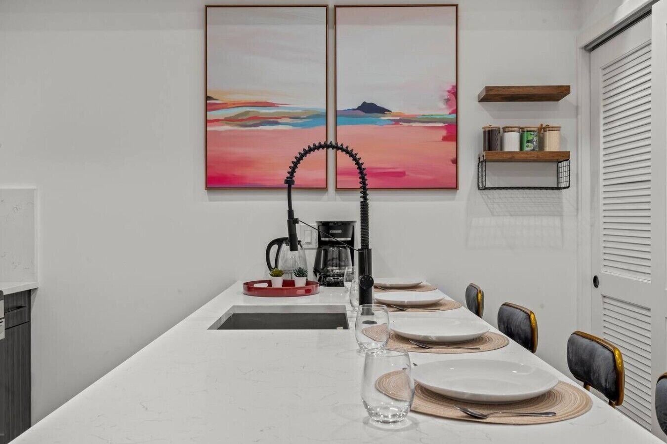 Een eethoek met een lange tafel gedekt voor vier personen, een koffieapparaat en twee abstracte schilderijen aan de muur.