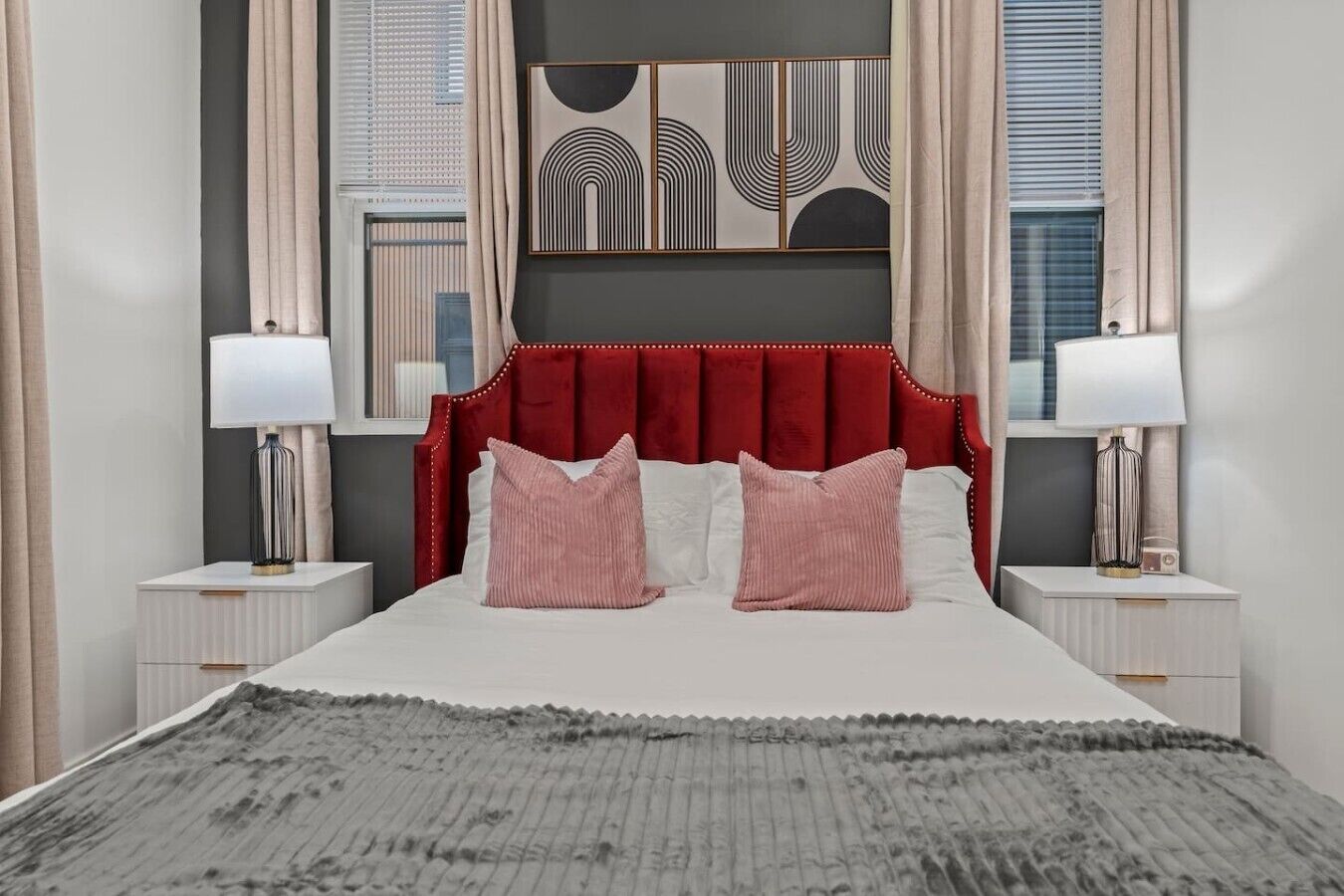 A bedroom with a red upholstered headboard, two bedside tables with lamps, a grey textured rug, and abstract wall art.