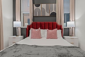A bedroom with a red upholstered headboard, two bedside tables with lamps, a grey textured rug, and abstract wall art. - 1BR | Art Museum, UPenn, University City, & Zoo (Philadelphia)