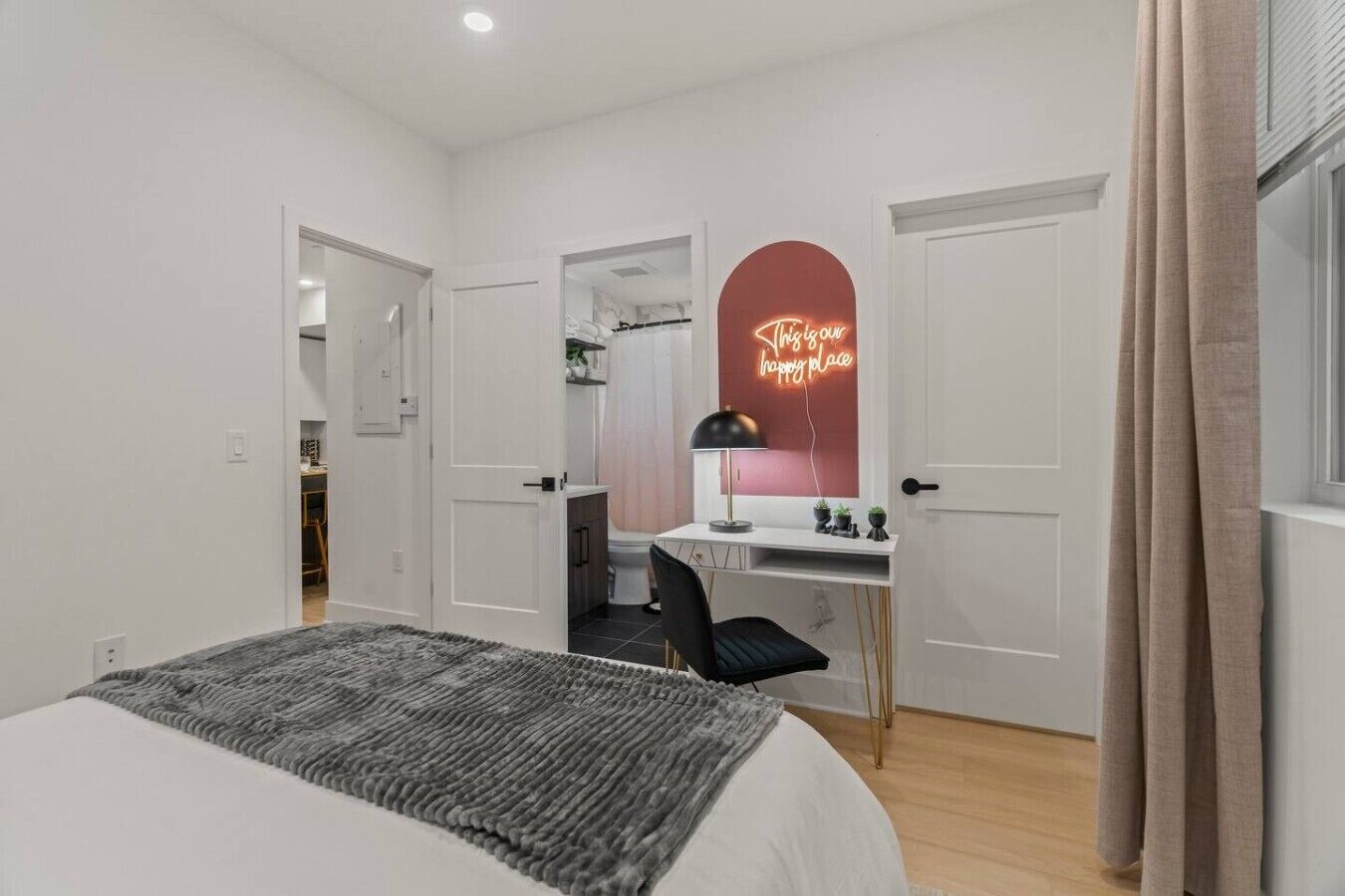A bedroom with a bed, a desk, a chair, a mirror, and a neon sign.