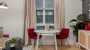 Ein Zimmer mit einem runden weißen Tisch, zwei roten Stühlen, einer weißen Kommode mit Korbmöbeln, einer großen Pflanze und einem Fenster mit beigen Vorhängen.
