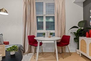 Ein Zimmer mit einem runden weißen Tisch, zwei roten Stühlen, einer weißen Kommode mit Korbmöbeln, einer großen Pflanze und einem Fenster mit beigen Vorhängen.
