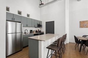 Cozinha moderna com armários cinza, geladeira de aço inoxidável, micro-ondas e cafeteira. Há uma pequena ilha com banquetas e uma mesa de jantar com cadeiras.