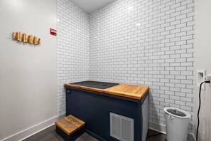 Canto de um quarto com parede de tijolos brancos, um pequeno banco de madeira e um eletrodoméstico preto com tampo de madeira.