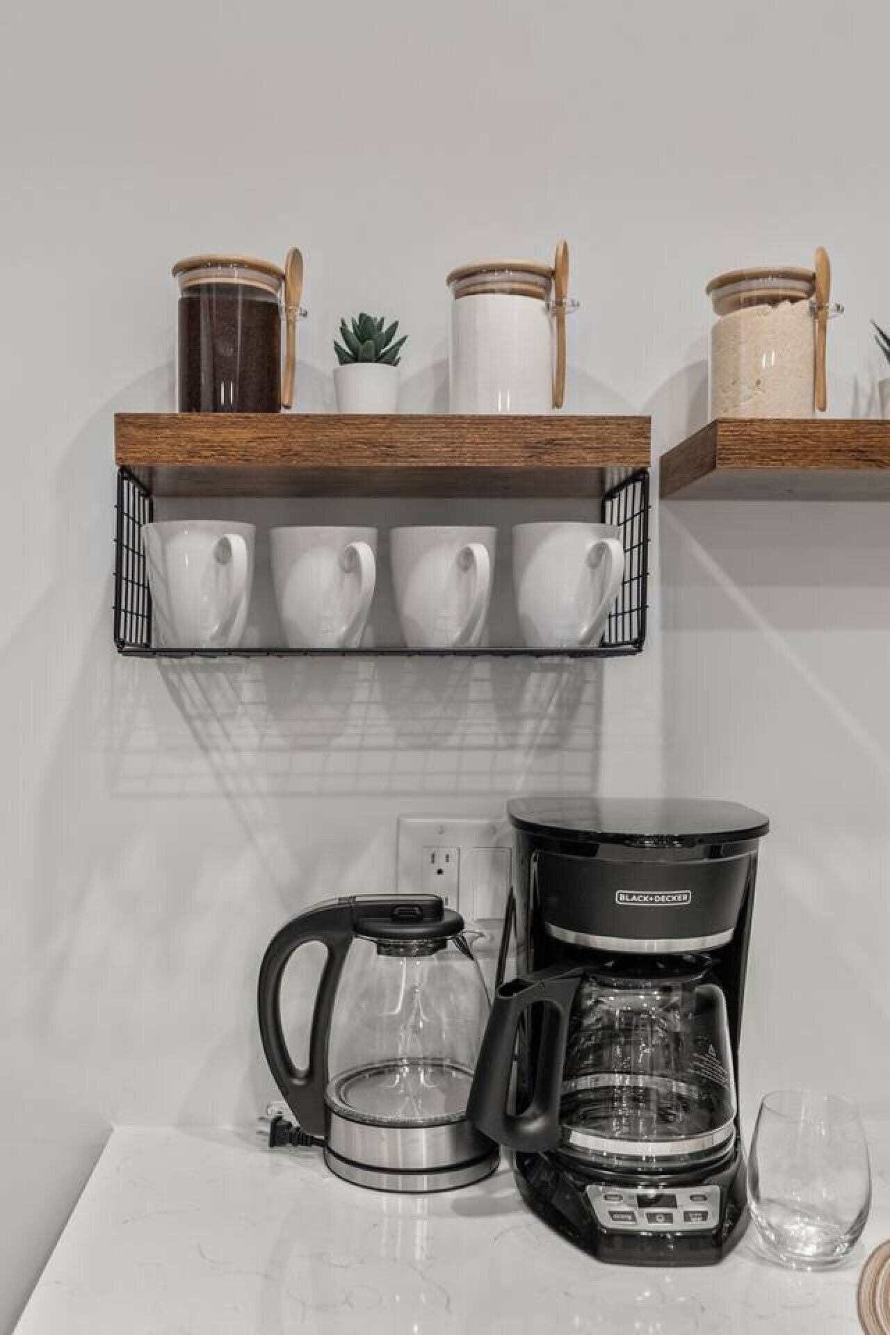 A kitchen counter with a coffee maker, a glass carafe, and a set of white mugs on a shelf.