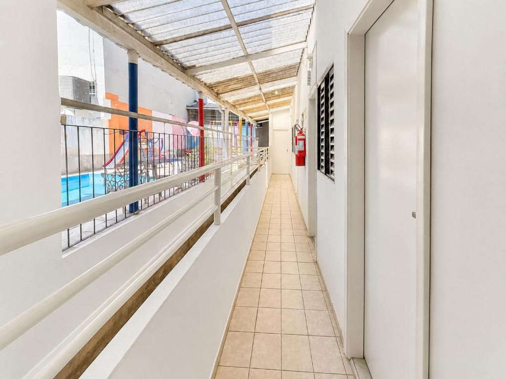 Corredor com piso de azulejos, corrimão de metal e vista para um parquinho infantil com um escorrega.
