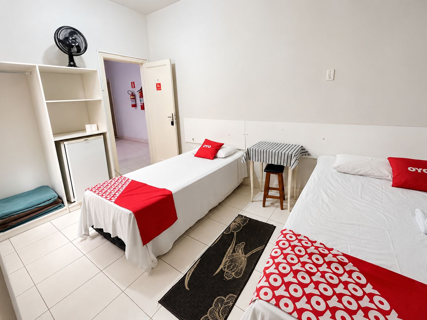 Een hotelkamer met twee bedden, een tafeltje, een krukje, een ventilator en een deur naar een badkamer.