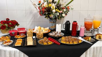 Mesa de buffet com diversos alimentos, um vaso de flores e dispensadores de bebidas.