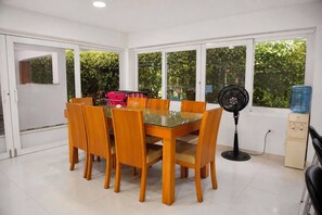 A dining area with a glass-top table, wooden chairs, a black standing fan, and a water dispenser.