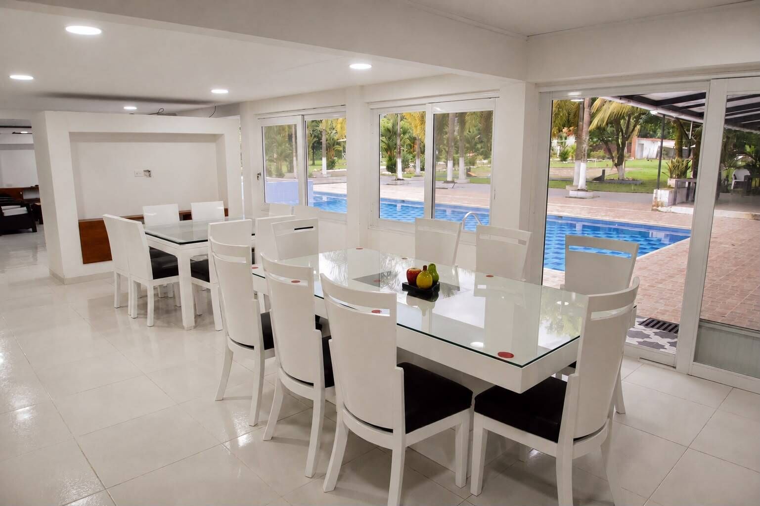 Un comedor con mesa de vidrio y sillas blancas, ventanas amplias con vista a una piscina y palmeras, y piso de baldosas.