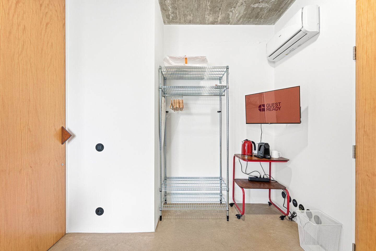 A hotel room with a metal rack, a red cart with a kettle and mug, and a 'GUEST READY' sign on the wall.