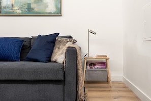 A living room with a grey sofa, a blue pillow, a patterned throw, a small wooden side table with a lamp, and a framed picture on the wall. - GuestReady - Fantastic Porto Flat (Porto)