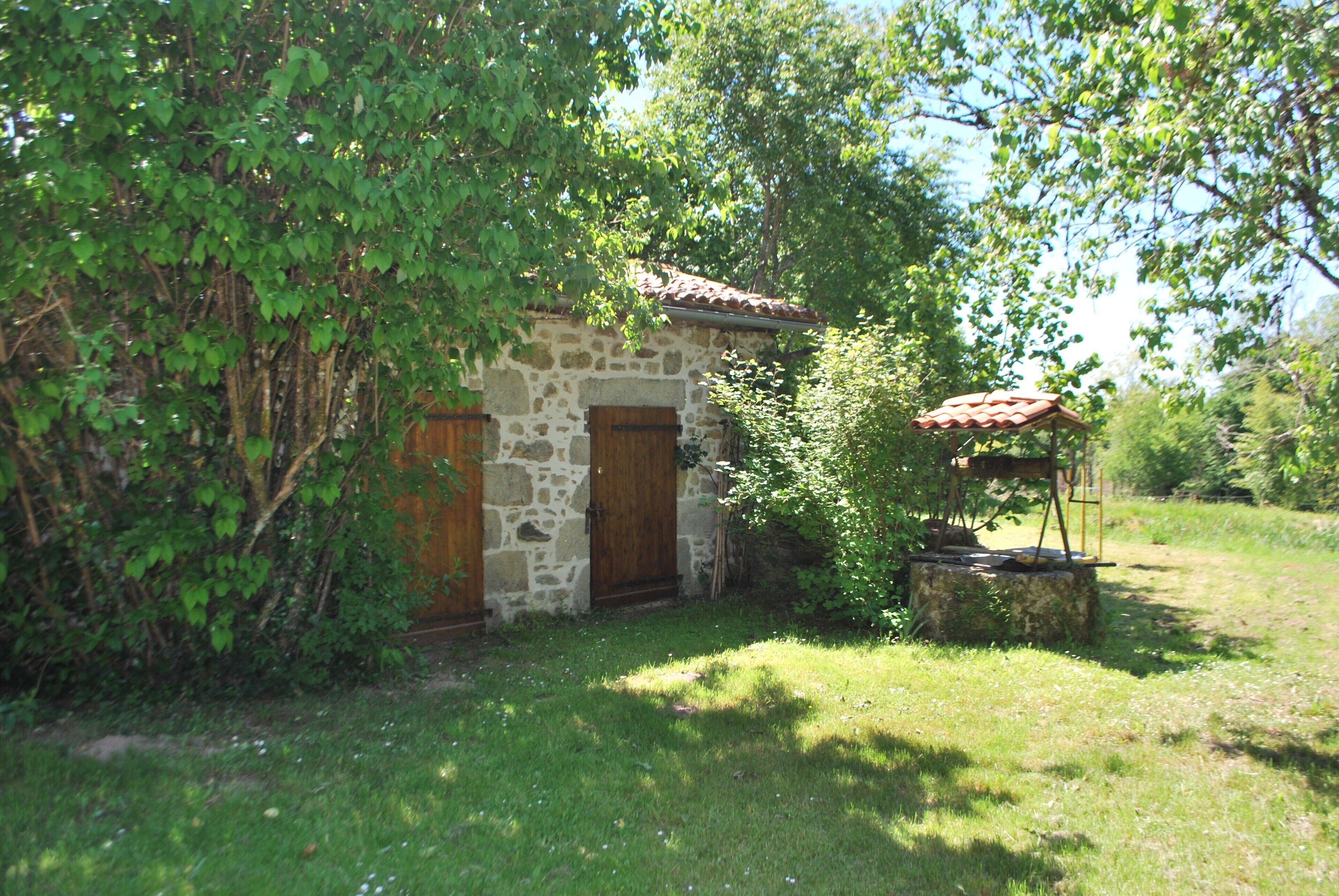 Un bâtiment en pierre avec une porte en bois, entouré de verdure et doté d’un petit espace extérieur aménagé pour s’asseoir.