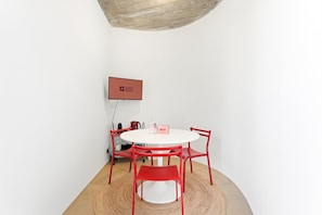 A room with a white round table, red chairs, a small rug, a flat-screen TV on the wall, and a red sign with 'GUEST READY' on it. - GuestReady - House of Many Faces - Room 3 (Porto)
