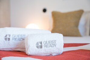 Two white towels with 'GUEST READY' text on a bed with a red blanket. - GuestReady - House of Many Faces - Room 3 (Porto)