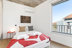 A hotel room with a bed, a small bedside table with a lamp, a window with a view of the outside, and an air conditioning unit on the wall. - GuestReady - House of Many Faces - Room 3 (Porto)