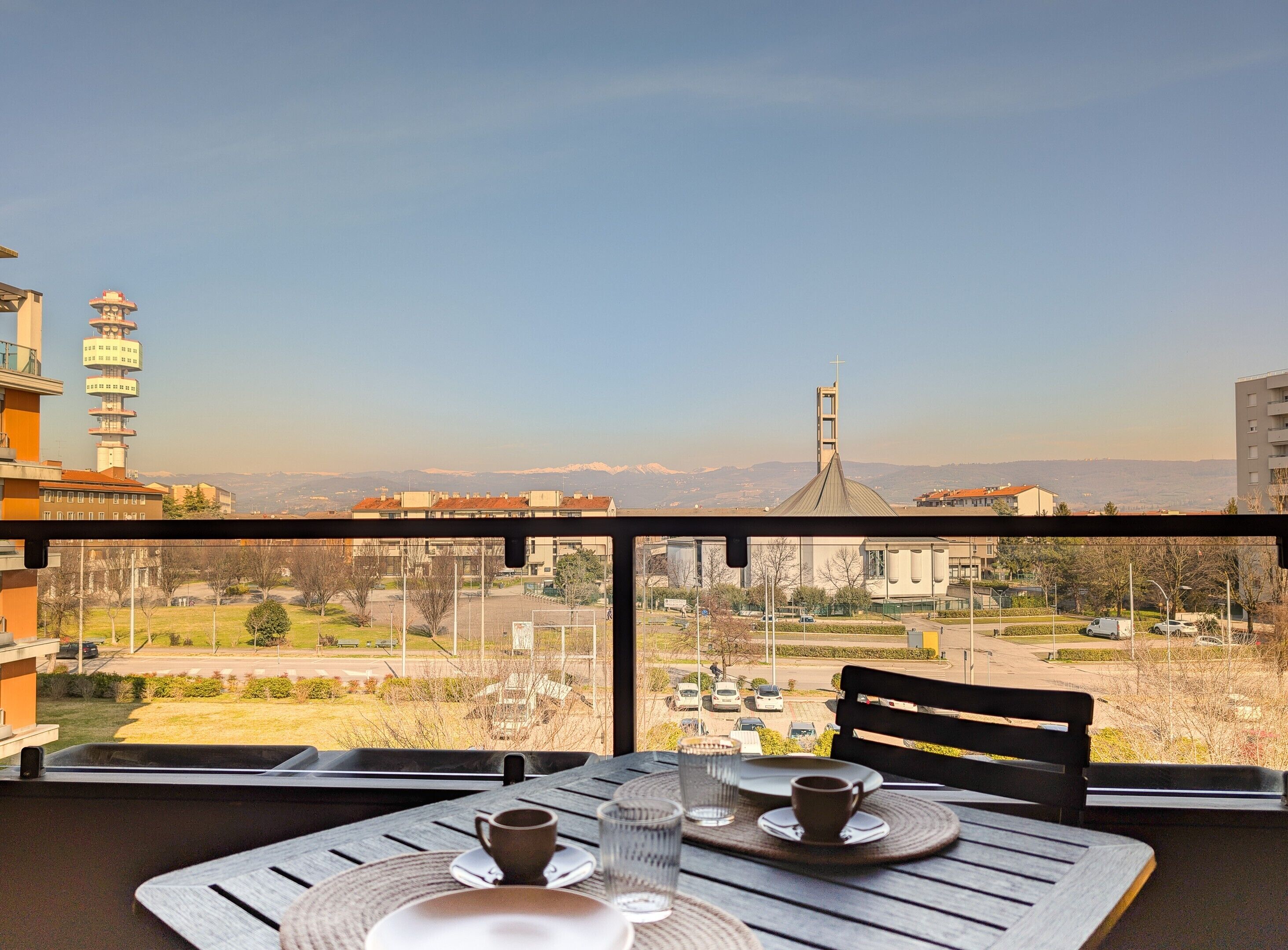 Un balcone con un tavolo apparecchiato per due, vista su un paesaggio urbano con edifici e montagne sullo sfondo.