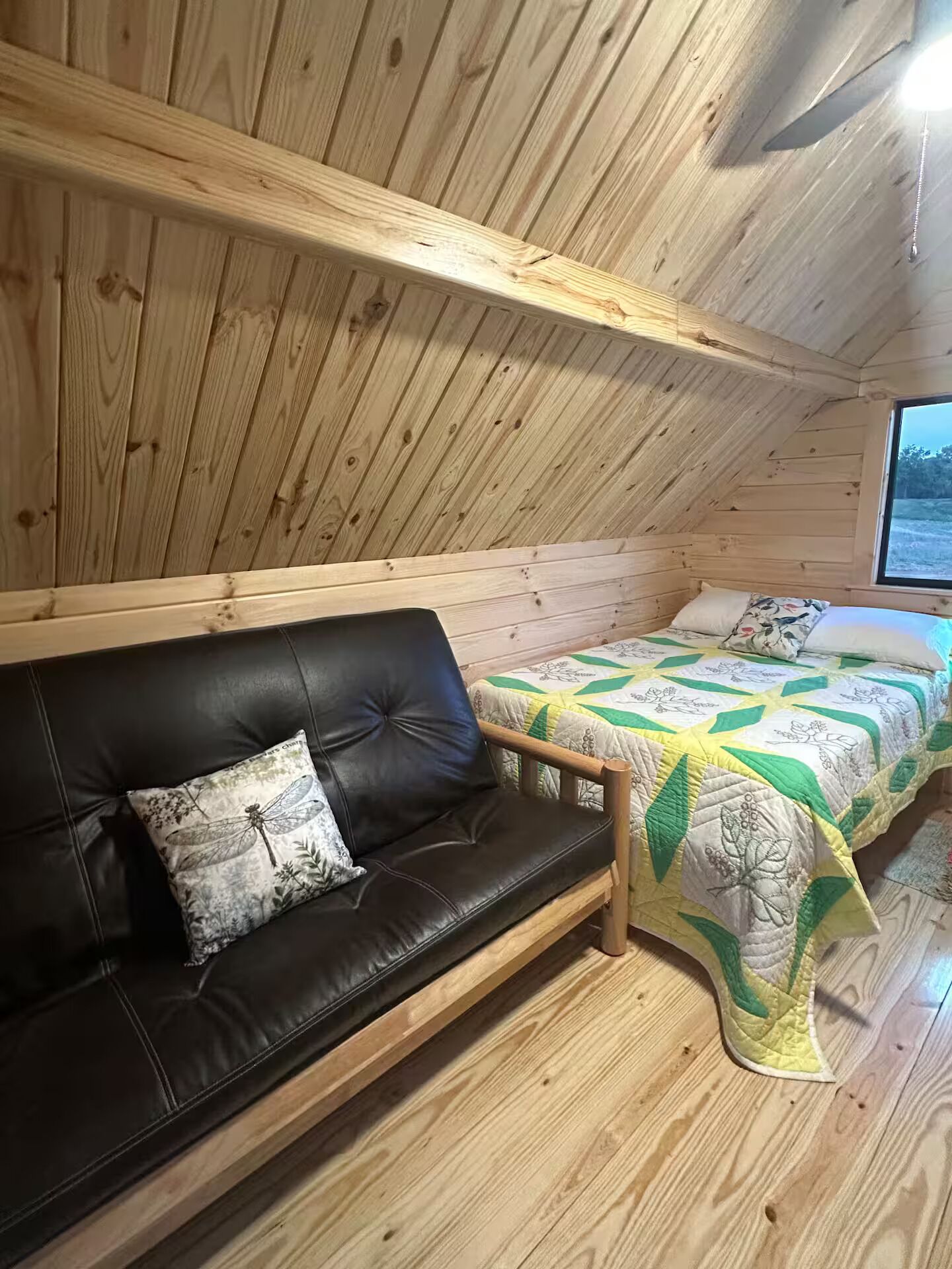 Interior de una cabaña con techo de madera, un sofá de cuero negro con un cojín floral y una cama con colcha verde y blanca.