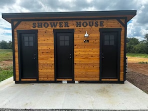 Een houten gebouw met de tekst 'SHOWER HOUSE', voorzien van drie zwarte deuren en een klein lampje boven het middenstuk.