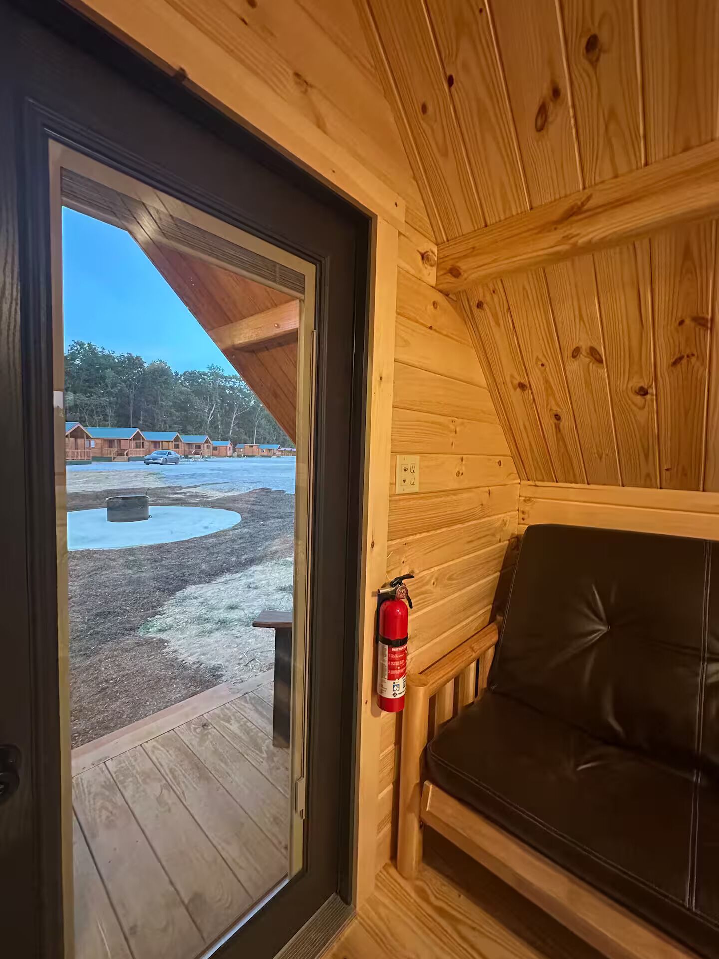 A wooden cabin interior with a door leading outside, a fire extinguisher mounted on the wall, and a black leather bench.