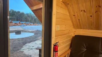 A wooden cabin interior with a door leading outside, a fire extinguisher mounted on the wall, and a black leather bench.