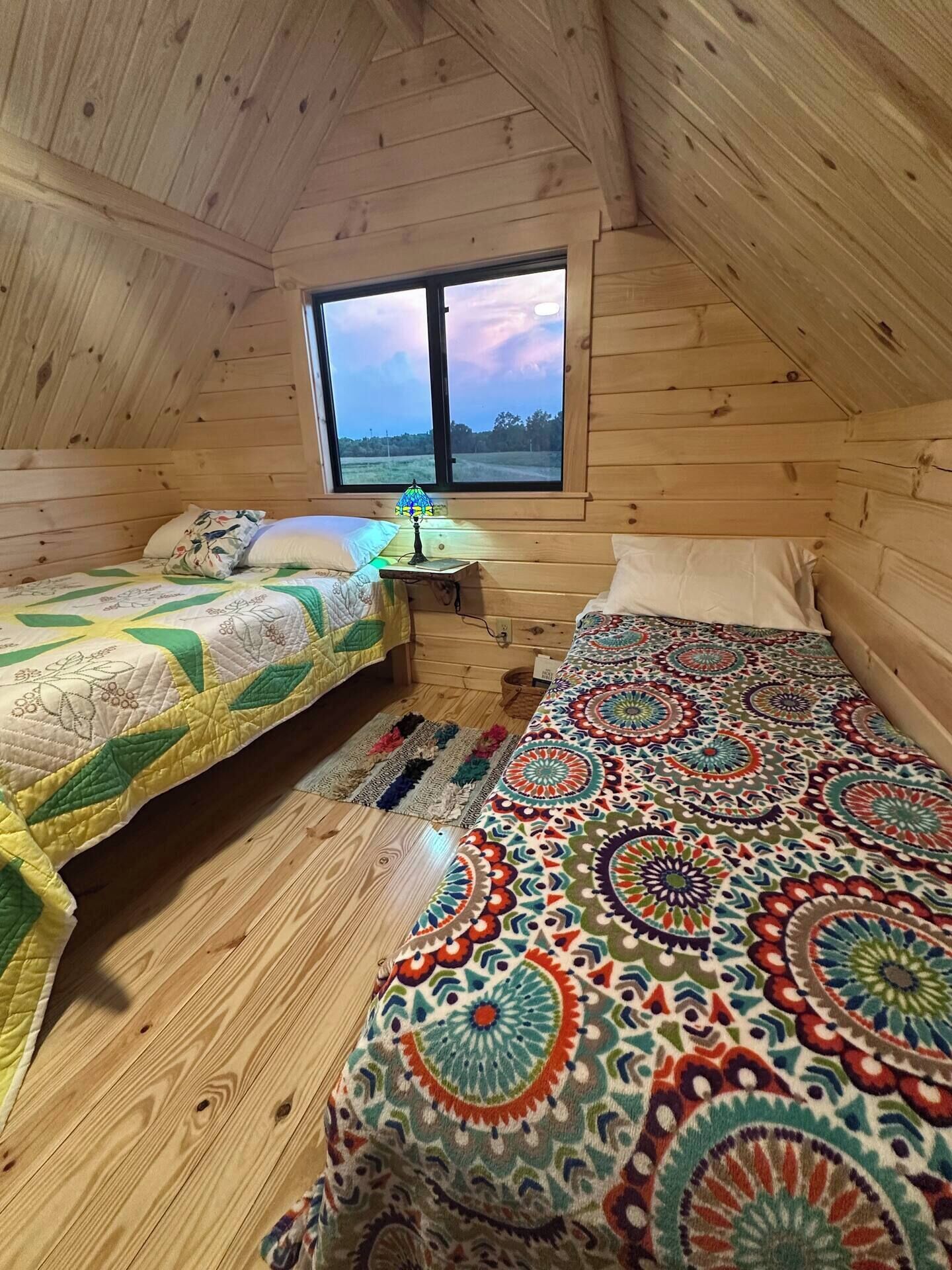 A cozy attic bedroom with wooden walls, a large window, two beds with colorful quilts, and a small table with a lamp.