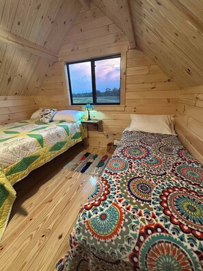 A cozy attic bedroom with wooden walls, a large window, two beds with colorful quilts, and a small table with a lamp.
