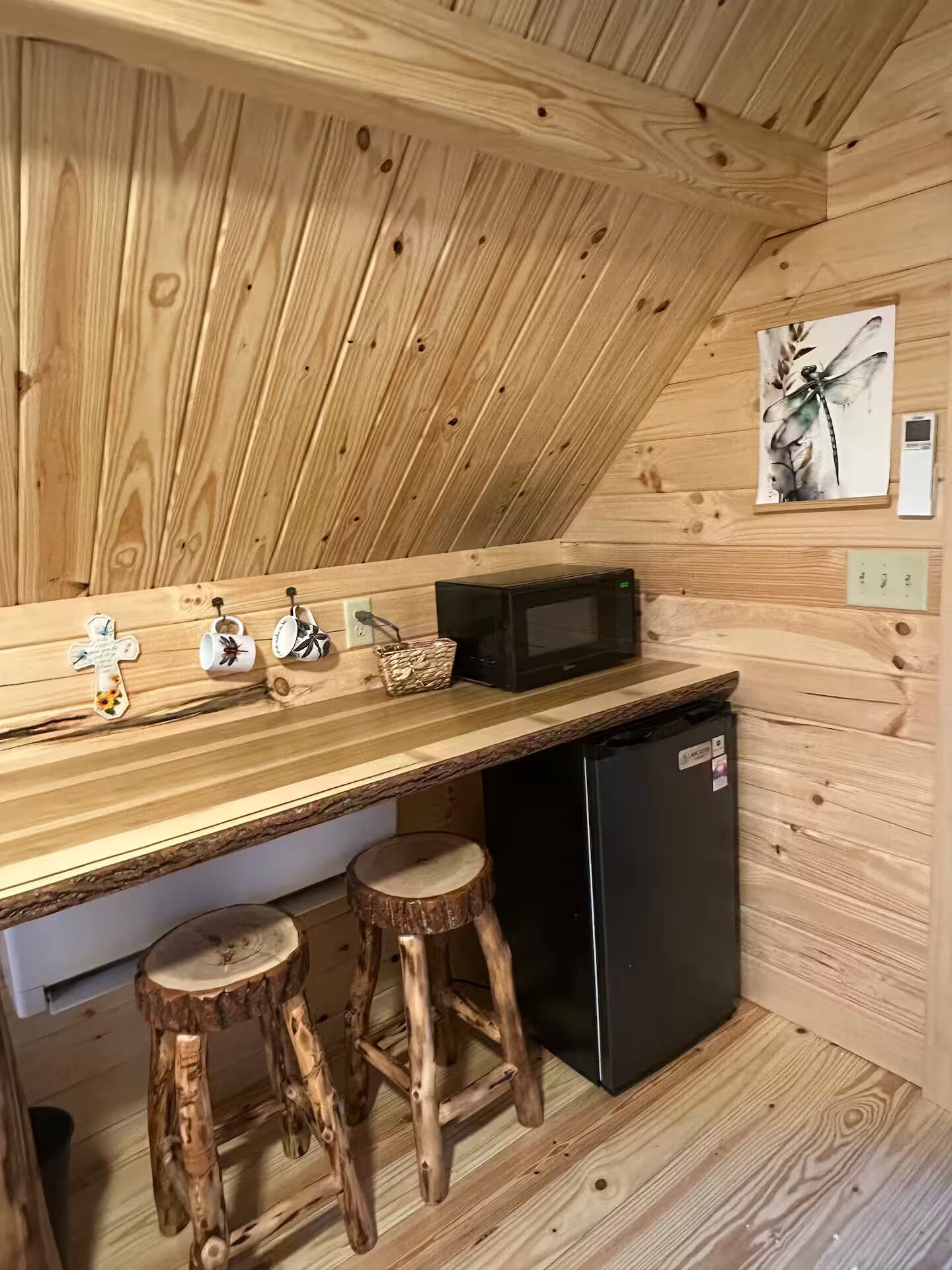 A wooden interior with a kitchen area featuring a microwave, a refrigerator, wooden stools, and a wall-mounted shelf with various items.