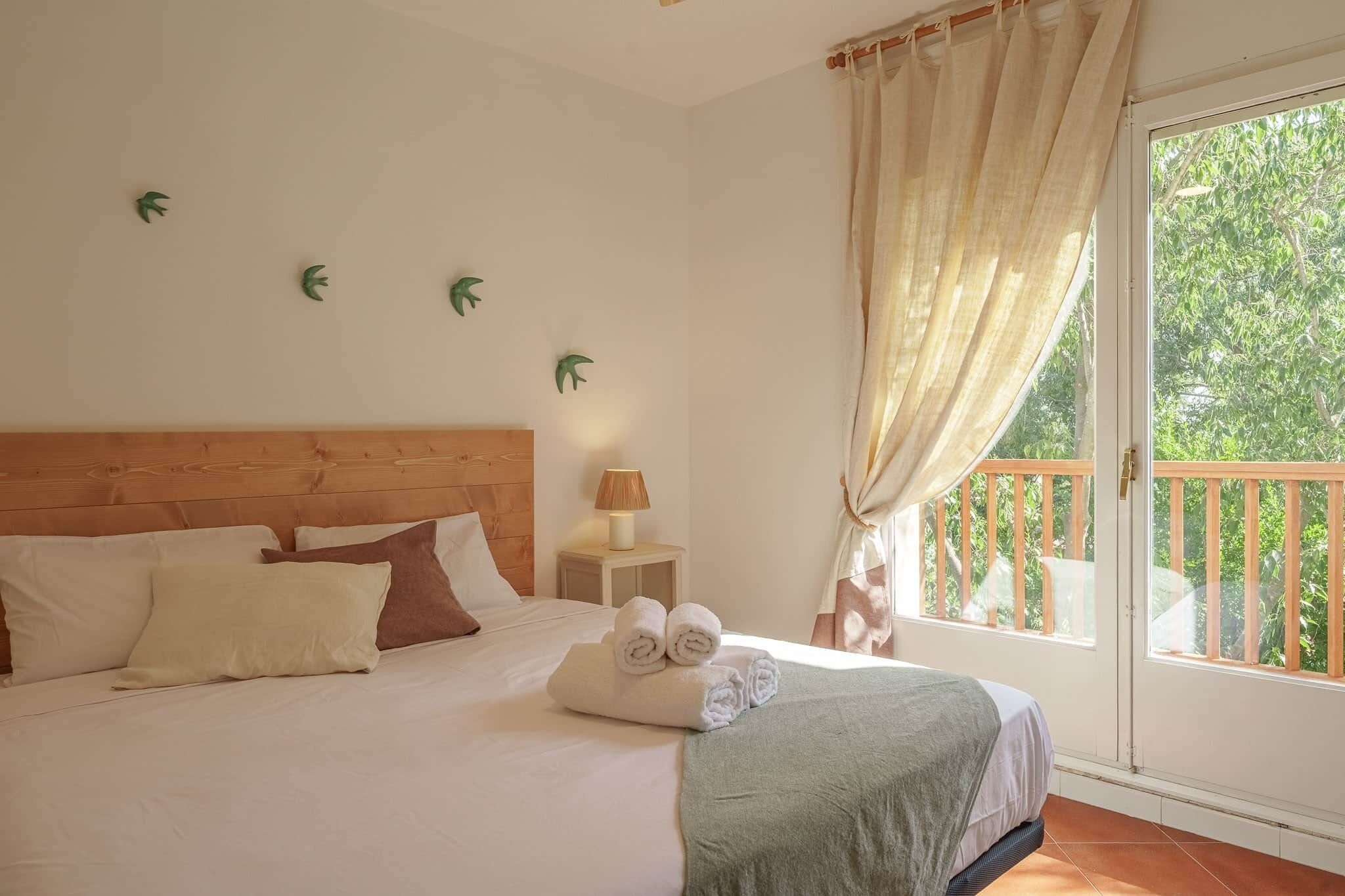 A bedroom with a wooden headboard, a bed with white bedding, a nightstand with a lamp, and a view of greenery through a balcony door.