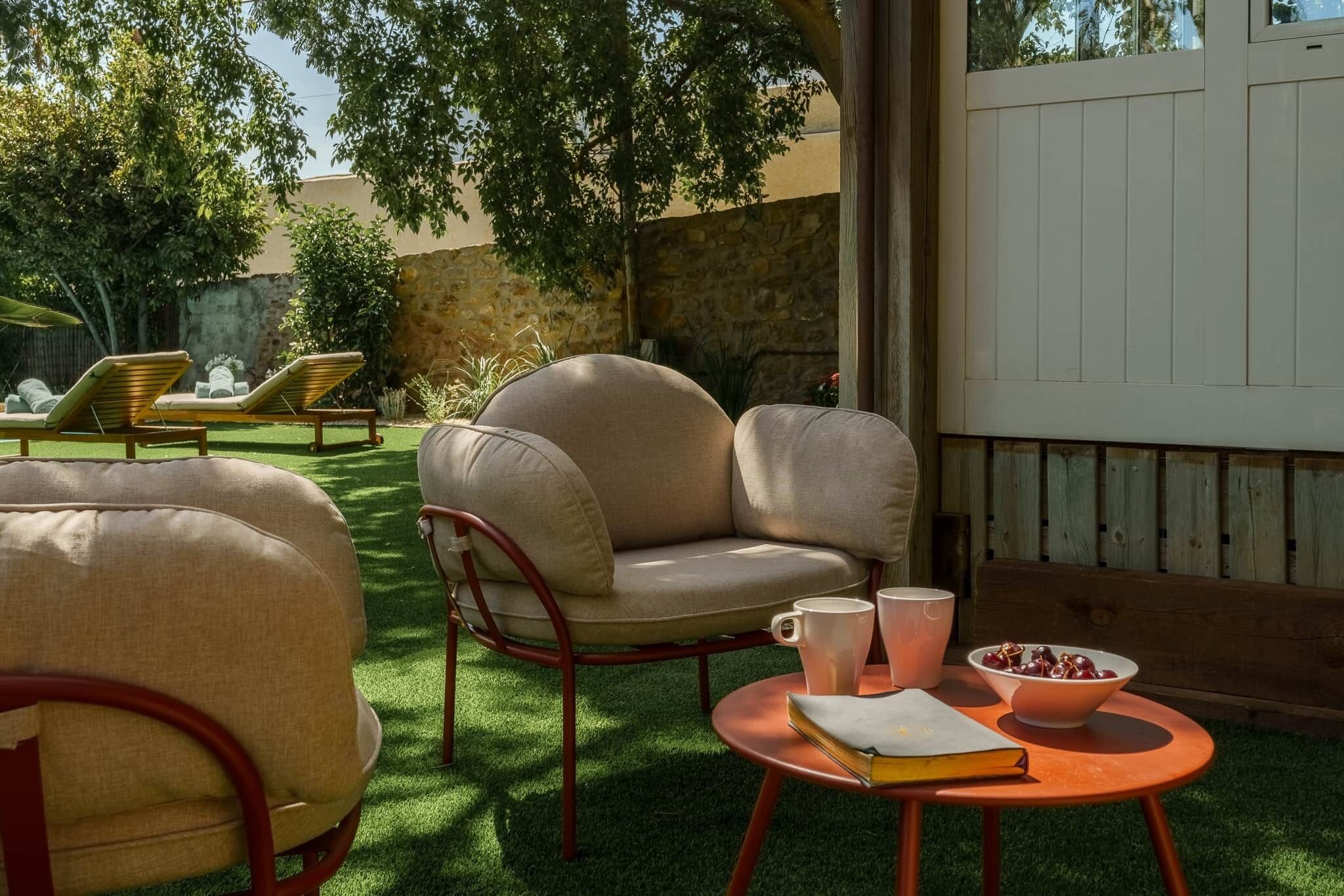 An outdoor seating area with cushioned chairs and a table, surrounded by greenery and a stone wall.